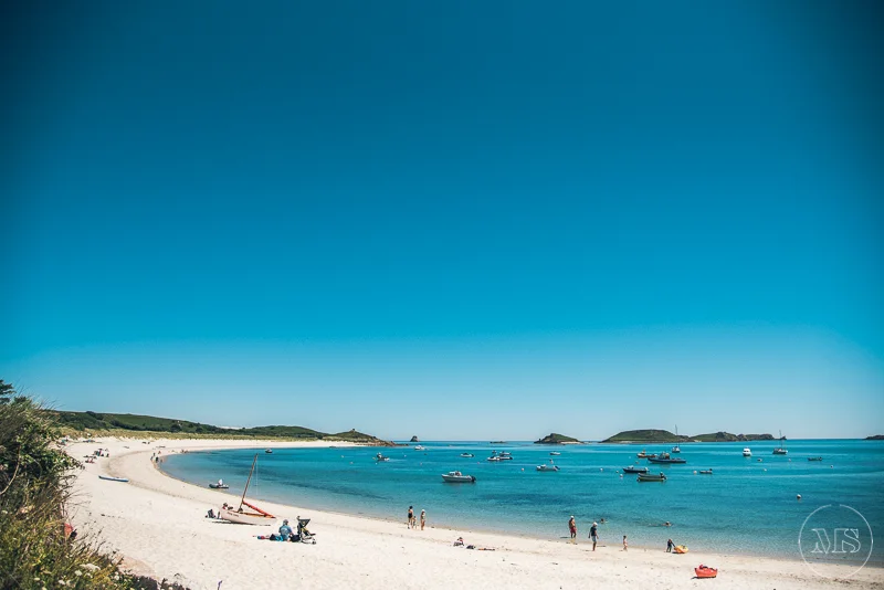 Isles of scilly photography. Sunny beach with boats on the water, people walking on the sand, and lush greenery on the side.