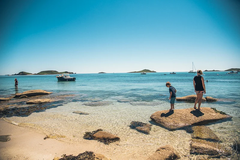 Isles of scilly photography. People enjoying a sunny day at the beach with rocks and boats in the water, and small islands in the background.