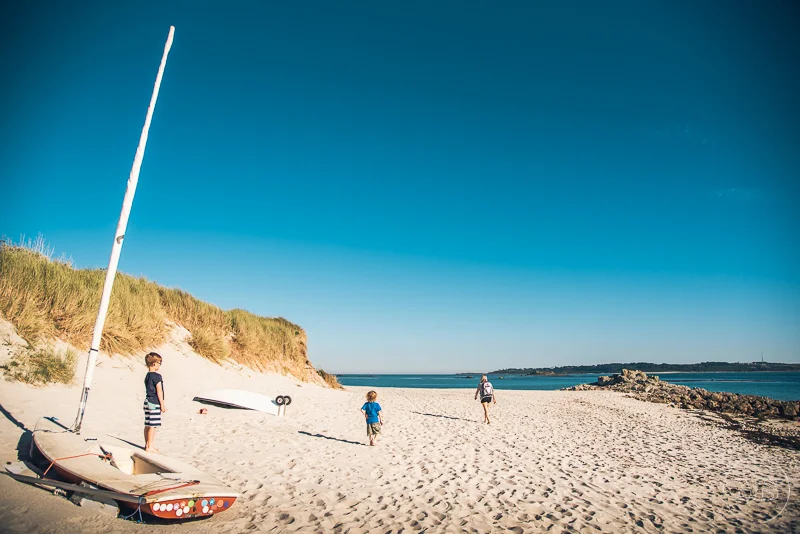 Beach scene with children and sailboat on sandy shore, grassy dunes, calm water, and clear blue sky.