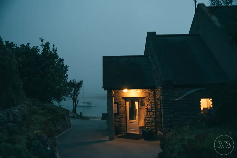 Isles of scilly photography. A stone house with warm lighting at night, surrounded by trees and overlooking water in the distance.