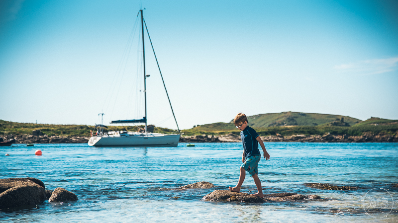 Young boy walking on rocks in shallow water at a beach with sailboats and grassy hills in the background - Isles of Scilly Professional Photographer - Mark Shaw Photography 