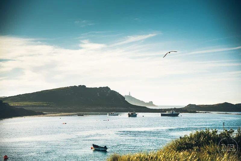 Isles of scilly photography. A tranquil bay with small boats floating on calm water, green hills surrounding the bay, a seagull flying in the sky, and a lighthouse on a distant hill under a partly cloudy sky.