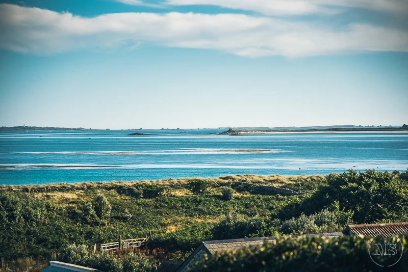 Isles of scilly photography. View of a coastal landscape with a large body of water, green vegetation, and some rooftops in the foreground, under a partly cloudy sky.