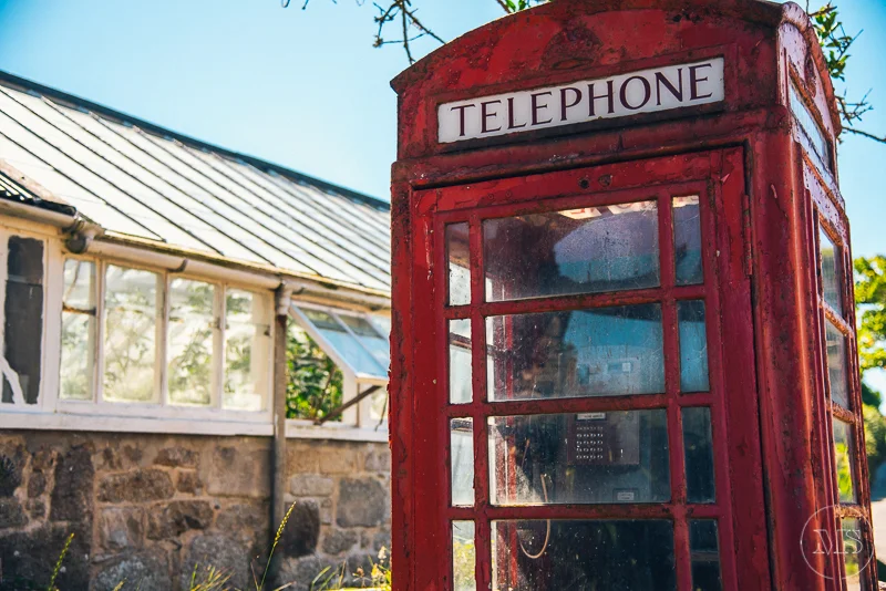 Close-up of a vintage red telephone booth outdoors with a stone building and clear blue sky in the background.
