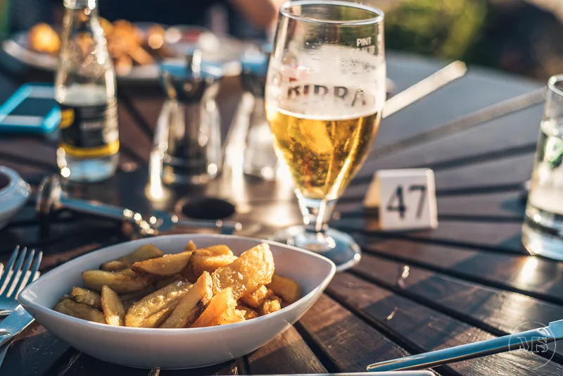 A bowl of French fries, a glass of beer, and a glass of water on a dark wooden outdoor table with a table number 47, with other drinks and condiments in the background.