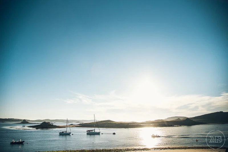 Isles of scilly photography. Calm body of water with several small boats anchored, rolling hills in the background, and a bright sun shining in the clear sky.