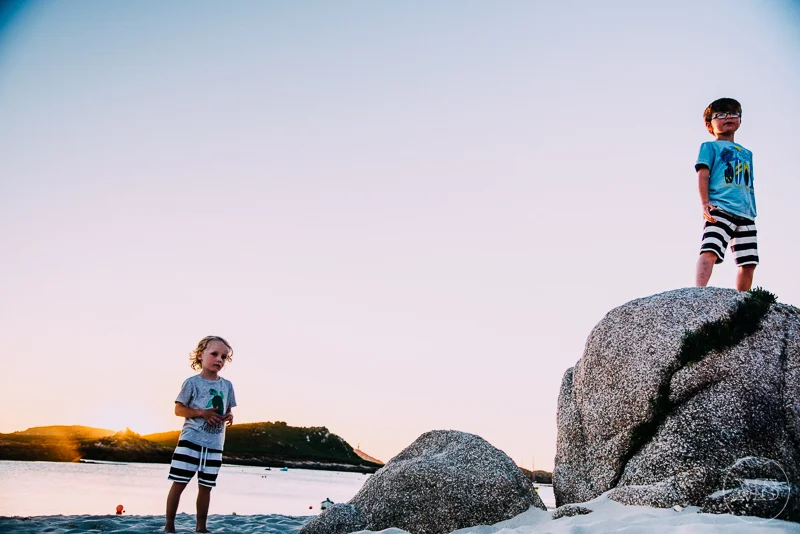Two young boys on a beach, one standing on large rocks and the other standing on the sand, with a water body and a hill in the background during sunset.