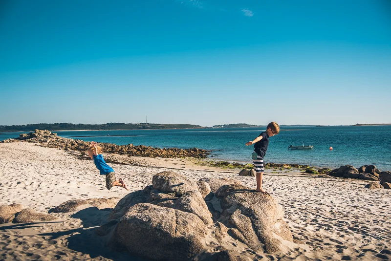 Two children playing on rocks and sand at a beach with calm blue water and a boat in the distance under a clear blue sky.