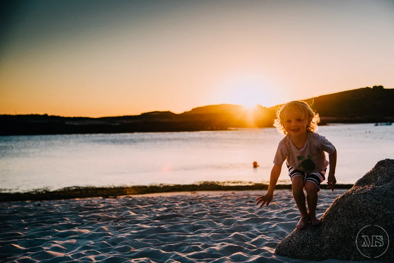 A young girl smiling and climbing on a rock on the beach at sunset, with a calm body of water and hilly landscape in the background.