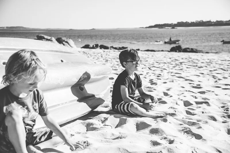 Two children sitting on a sandy beach near a kayak, with the ocean and boats in the background, black and white photo.