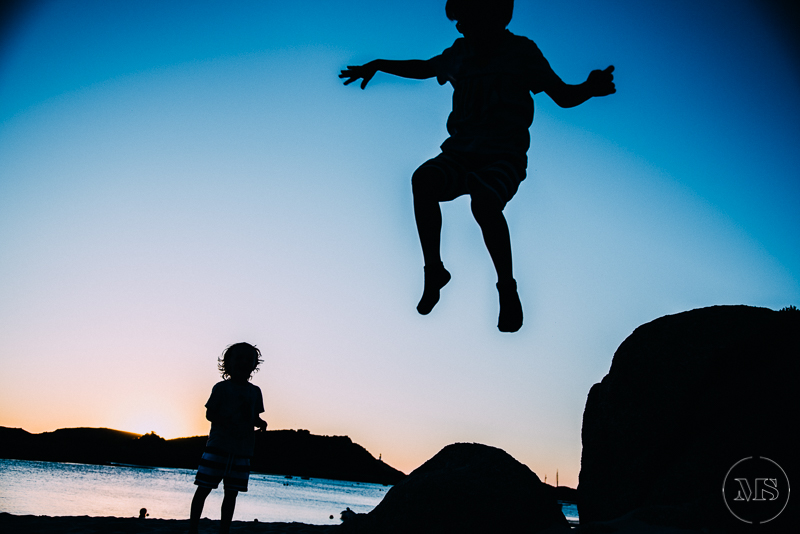 Silhouette of a person jumping in the air with arms outstretched, next to a child standing on rocks at sunset by the water - Isles of Scilly Professional Photographer - Mark Shaw Photography 