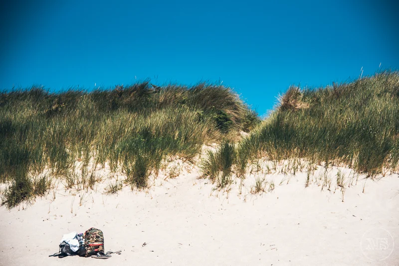 Isles of scilly photography. Sand dune on a beach with tall grass and a blue sky, a backpack and a towel on the sand in the foreground.