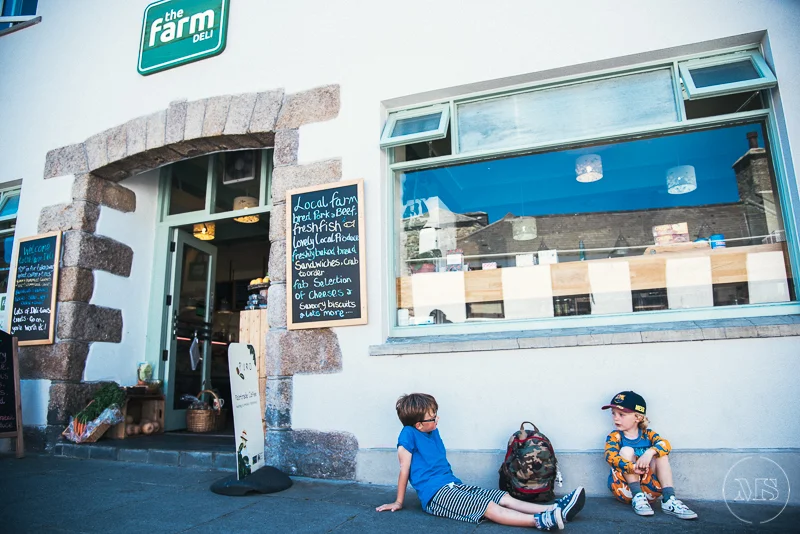 Children sitting outside the entrance of a farm shop, with a chalkboard menu and large window showing the interior.