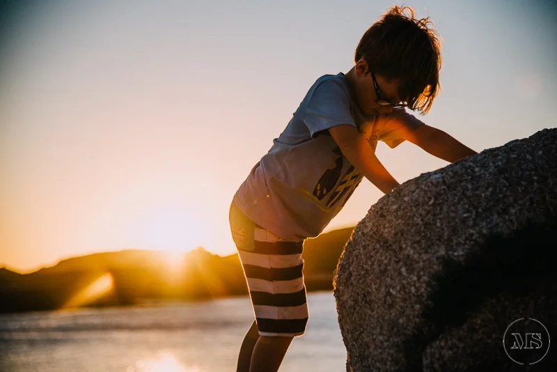 Isles of scilly photography. Child wearing glasses and striped shorts leaning on a large rock at sunset by the water.