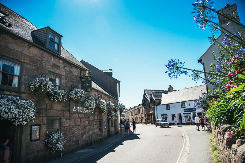 Isles of scilly photography. A sunny street scene with historic stone buildings adorned with flower boxes, pedestrians, and parked cars under a clear blue sky.