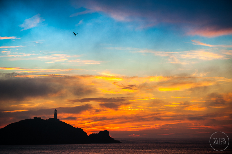 Isles of scilly photography. A sunset over the ocean with an orange and yellow sky, silhouette of a small island with a lighthouse, and a bird flying in the sky.