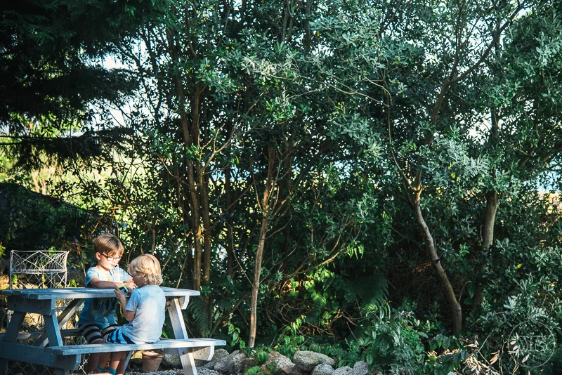 Isles of scilly photography. Two children sitting at a picnic table in a lush, green outdoor setting, possibly a backyard or park, surrounded by trees and foliage.