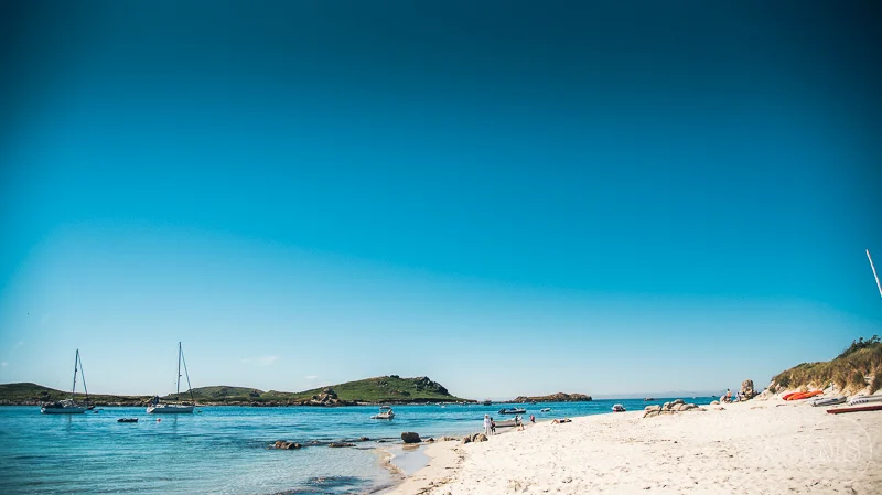 Sandy beach with sailboats anchored in calm water and a grassy island in the background under a clear blue sky - Isles of Scilly Professional Photographer - Mark Shaw Photography 