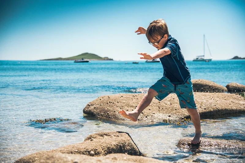 Isles of scilly photography. Young boy jumping between rocks on a beach near the water, with boats and islands in the background on a sunny day.