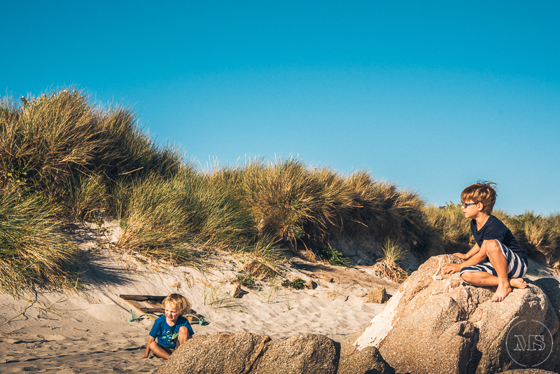 Two children playing on beach sand with large rocks, grassy dunes in the background, under a clear blue sky.