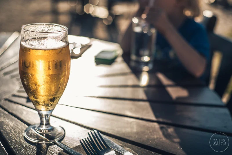 A glass of beer on a wooden outdoor table with a blurred background of a person and another glass.
