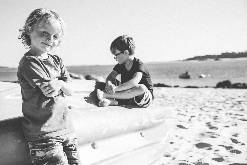 Two children on a beach; one is standing with arms crossed and the other is sitting on a boat, writing in a notebook.