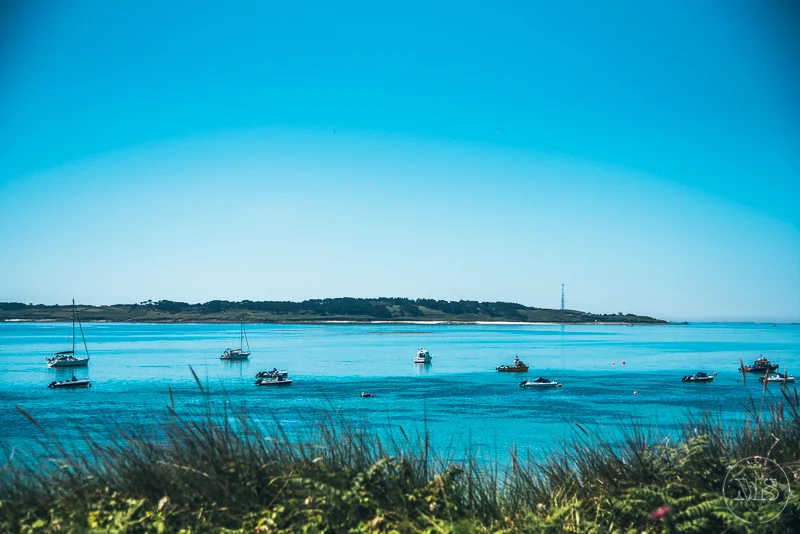 Isles of scilly photography. Calm bay with sailboats and motorboats, green shoreline in background under clear blue sky.
