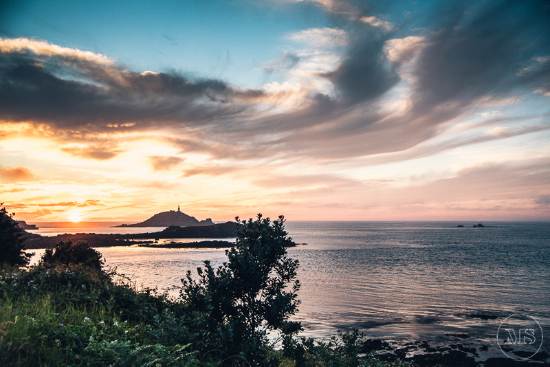 Sunset over the ocean with clouds and a hill in the distance, framed by greenery in the foreground.