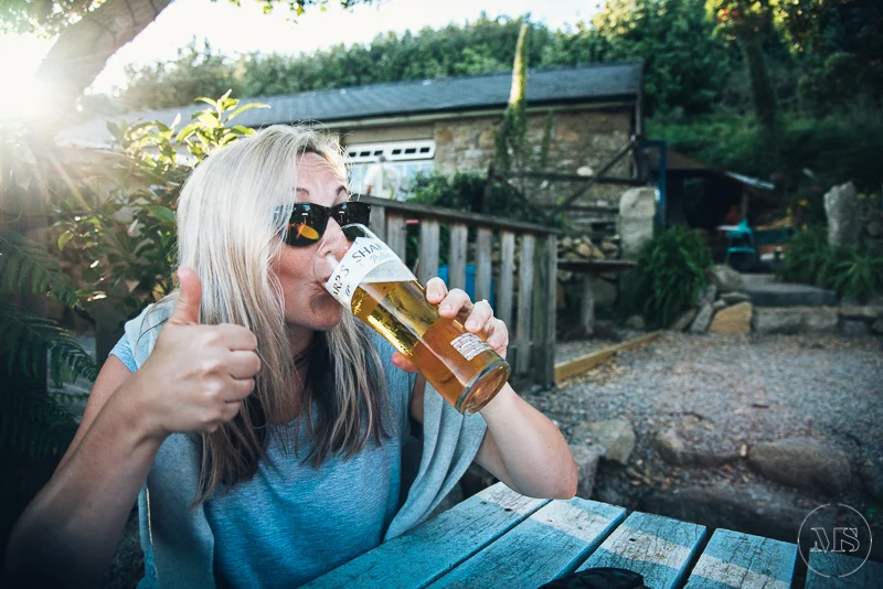 Isles of scilly photography. A woman with long blond hair, wearing sunglasses and a gray shirt, is enjoying a glass of beer outdoors, giving a thumbs-up sign.