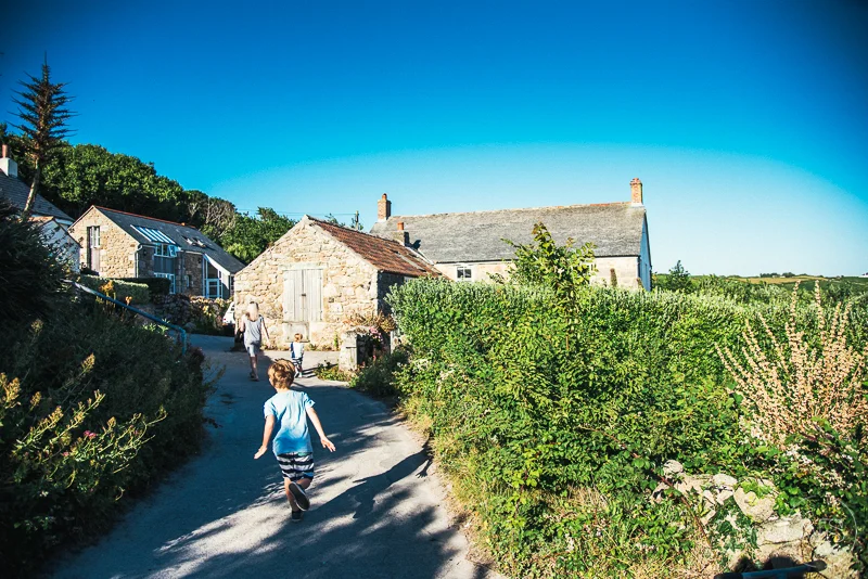 Children walking down a country road past stone houses and lush greenery under a clear blue sky.
