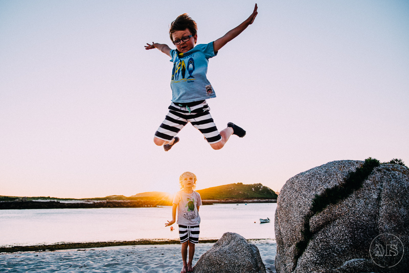 Two children on the beach at sunset, one jumping in the air with arms outstretched and the other standing on the sand, with large rocks nearby and a boat in the water.