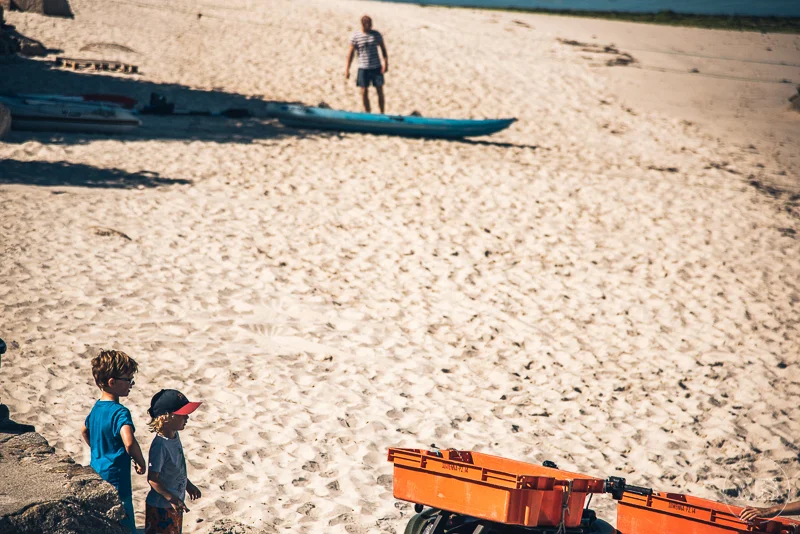 Two young boys standing on a sandy beach, looking toward a person walking in the distance near the water, with a kayak and equipment nearby.