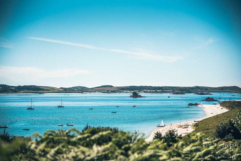 Isles of scilly photography. A scenic view of a bay with sailboats and small boats, sandy beaches, green hills, and a bright blue sky.