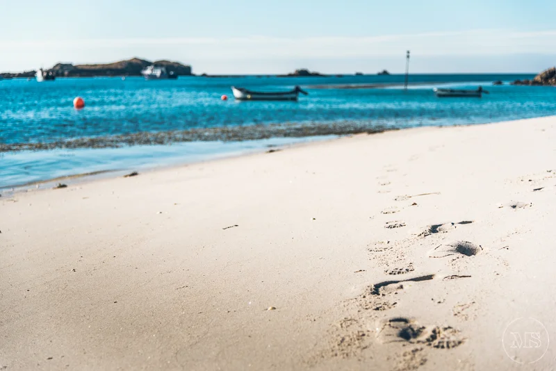 Footprints on a sandy beach near calm blue water with boats anchored, and a small island or landmass in the distance under a partly cloudy sky.