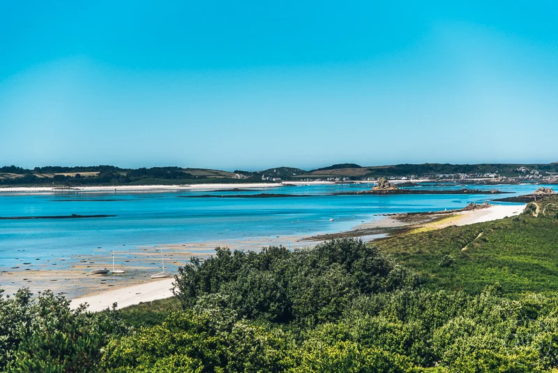 A wide view of a coastal landscape with blue water, sandy beaches, green vegetation, and a distant shoreline under a clear blue sky.