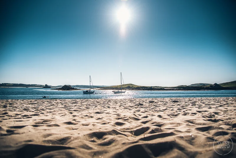 Sandy beach with two sailboats anchored in a calm bay, hills in the background, bright sun in a clear blue sky.