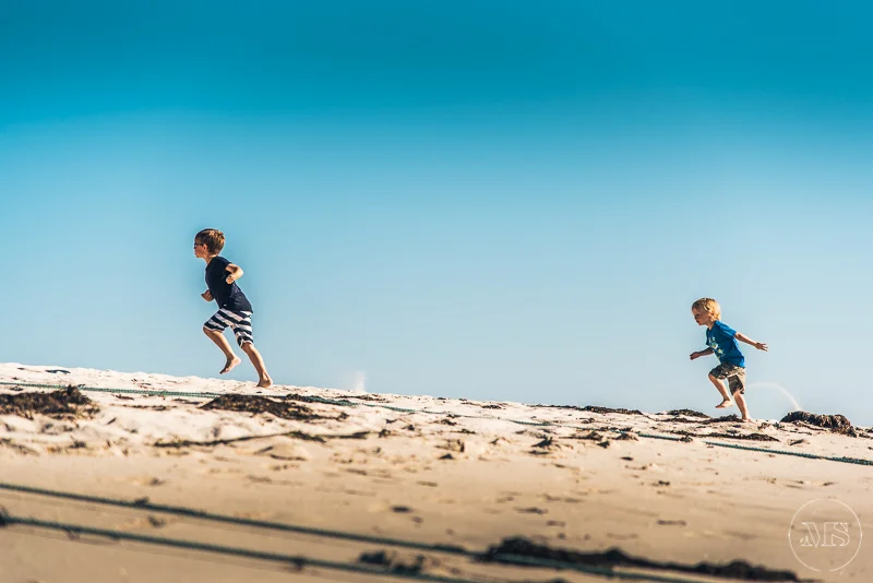 Two boys running and playing on the sandy beach under a clear blue sky.