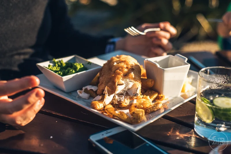 Plate with breaded fish fillet, French fries, green salad, and two small white sauce containers, set on a dark wooden table beside a glass of water with cucumbers and a smartphone. Two people are visible, one holding a fork and the other using a phon