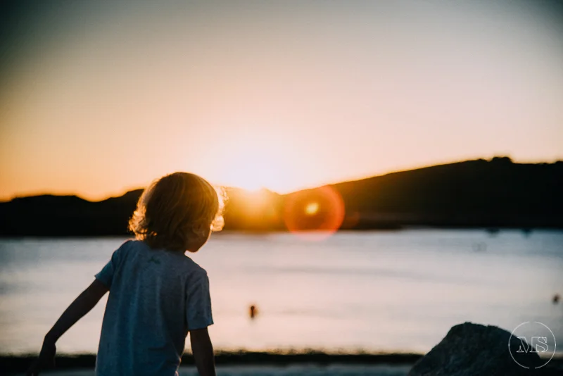Child sitting on rocks by the water during a sunset or sunrise.