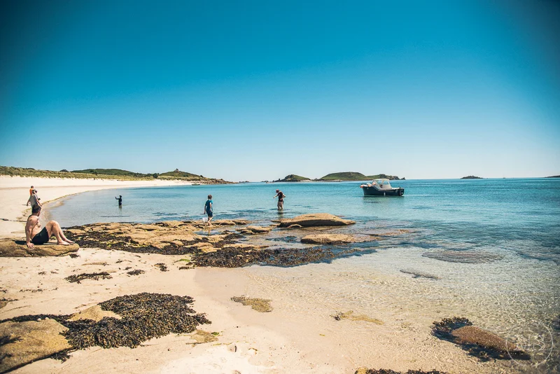 People relaxing and swimming on a sandy beach with rocks, clear blue water, and boats in the distance.