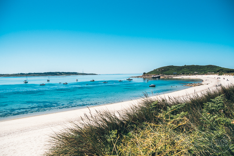 Isles of scilly photography. A beach scene with white sandy shore, clear blue water, boats anchored near the shore, and green hills in the background under a bright blue sky.
