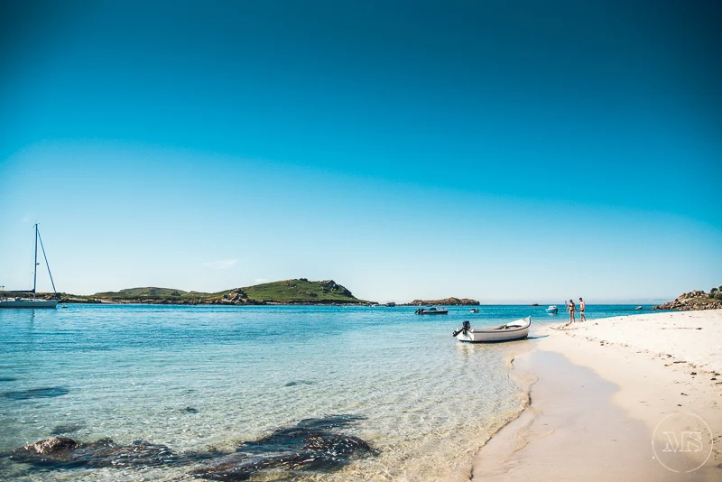 Isles of scilly photography. A beach scene with clear blue water, a white sandy shoreline, small boats anchored near the shore, and a few people standing on the beach under a bright blue sky.