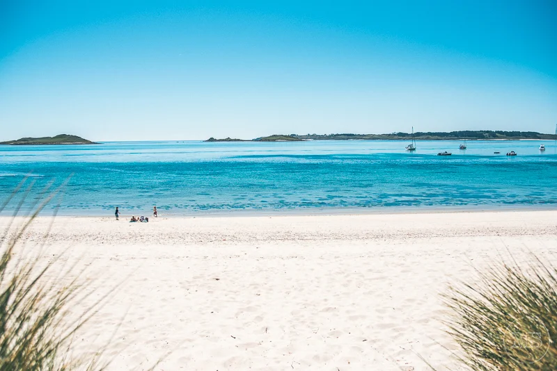 Isles of scilly photography. Beach with white sand, blue water, clear sky, and a few people enjoying the scenery, with sailboats anchored offshore.