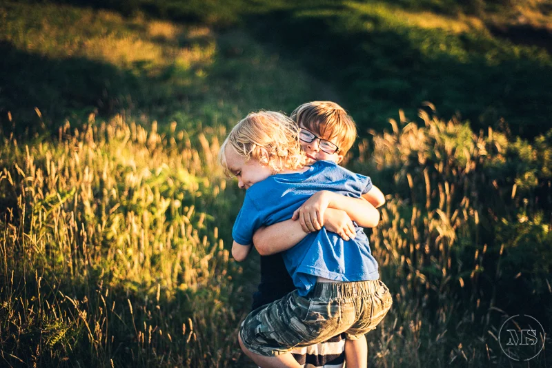 Two children, a boy with glasses and a girl, hugging and playing together outdoors in a grassy field during sunset.