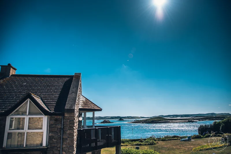 Sunny day view of a house with a stone exterior and a black roof overlooking a body of water with small islands in the distance.