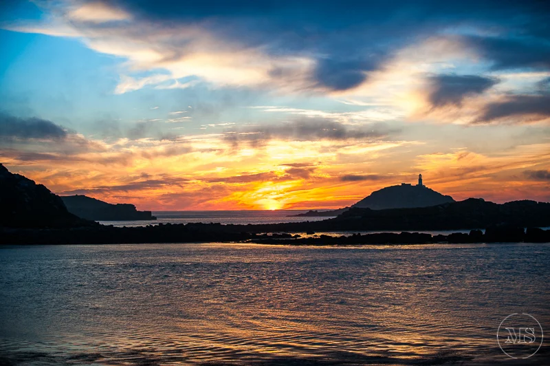 Sunset over a body of water with silhouettes of hills and a lighthouse on one hill.