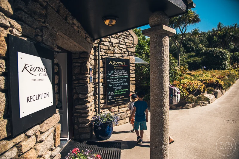People walking past the entrance of a building with stone walls, with signs for "Karma" and a reception area, near a landscaped garden with plants and a pathway under a sunny sky.