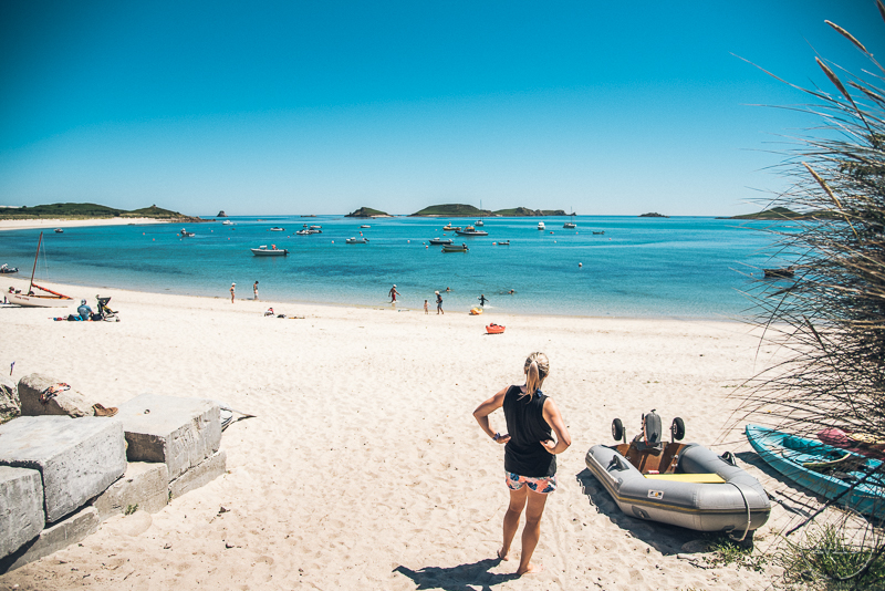 Isles of scilly photography. A woman stands on a sandy beach looking at boats in the water with small islands in the distance.