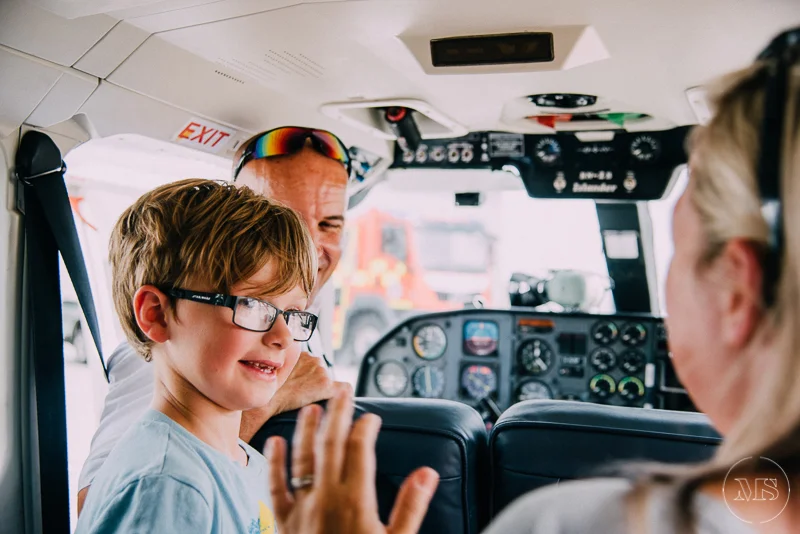 Isles of scilly photography. A young boy with glasses sitting in an airplane cockpit, smiling and looking at a woman, with an adult man smiling behind him. The woman appears to be explaining or demonstrating something.