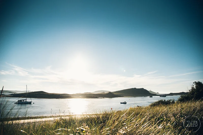 Isles of scilly photography. A serene coastal scene with boats anchored on calm water, grassy shoreline in the foreground, hills in the background, and a bright sky with the sun low on the horizon.